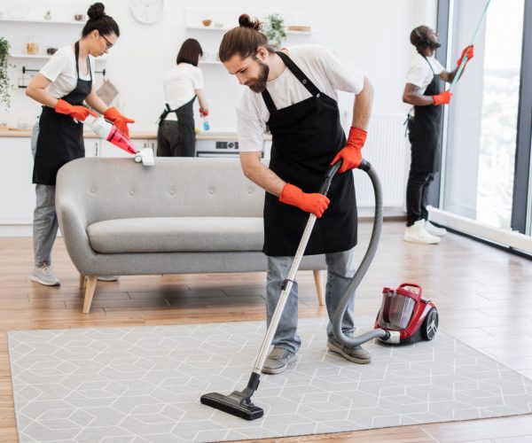 Team of professional people from cleaning service cleans sofa, carpet, cuisine table and panoramic windows of spacious apartment. Multinational group of cleaners cleans bright studio kitchen.